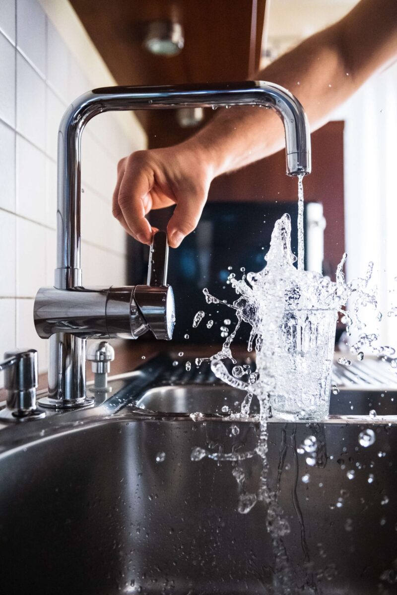 Image of a glass cup being filled with water from the sink; only an arm and hand are showing